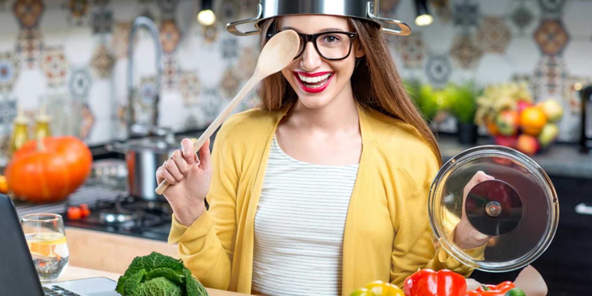 בחורה במטבח עם סיר על הראש מכסה את העין עם כף עץ ומחייכת